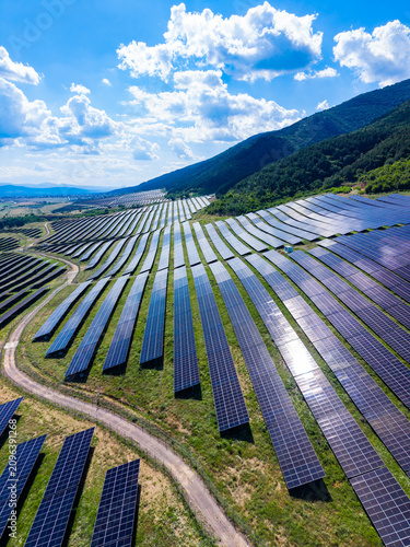 Solar panels stretch across a hillside in a green valley under a blue sky with fluffy clouds in the afternoon