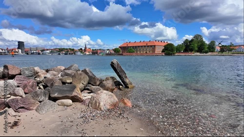 View across Flensburg Fjord toward the historic Sonderborg with clear water, a rocky shoreline, sandy beach and Mill in the background.