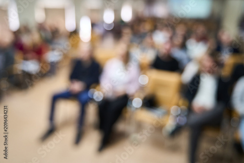 Audience Seated Listening Lecture Hall Conference Attendees In Auditorium Facing Forward During Corporate Seminar With Rows Of Tan Chairs And Soft Blurred Focus Capturing Group Attendance.