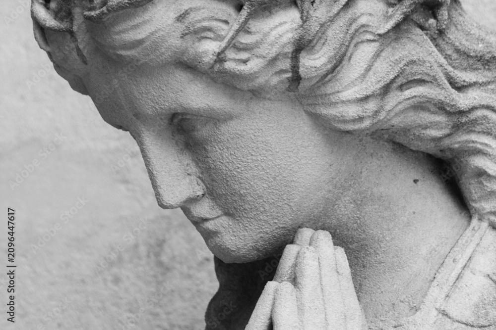 Fototapeta premium Black and white close-up of an ancient praying angel statue with folded hands. A timeless symbol of faith, hope, prayer, peace, divine protection and spiritual comfort.