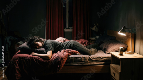 Woman Sleeping Exhausted In Dimly Lit Bedroom On A Messy Bed At Night