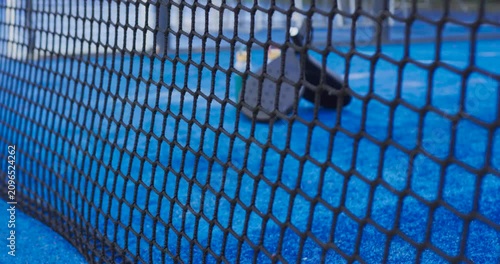 Camera is pulling back from extreme close-up, revealing padel rackets and bottle on blue court
