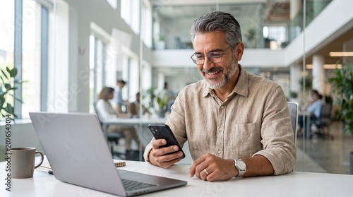 Middle aged Hispanic business manager ceo using cell phone mobile app, laptop. Smiling Latin or Indian mature man businessman holding smartphone sit in office working online on gadget with copy space.