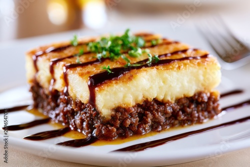 Plated shepherd's pie square with thick layers of mashed potatoes and ground meat, garnished with parsley, served on a white dish with sauce drizzled around