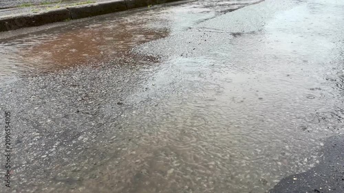 Rainwater accumulates on the asphalt surface, forming puddles as raindrops create ripples, highlighting a rainy urban street scene