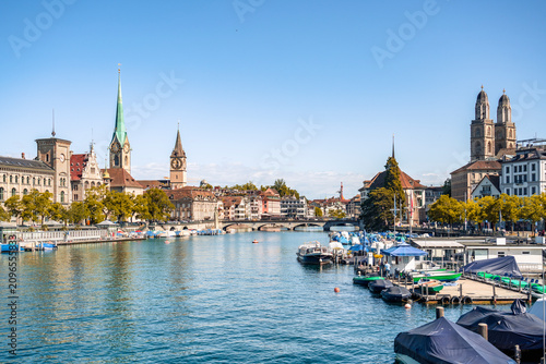 This picturesque scene captures the Limmat River flowing through the historic center of Zurich, Switzerland, framed by iconic church spires and docked boats.