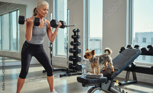 Senior Woman Lifting Dumbbells with Small Dog in Gym