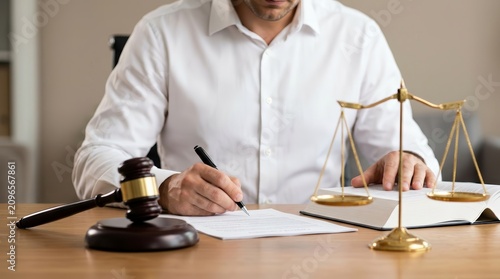 Male lawyer signing legal documents at a desk with a judge gavel, law book, and scales of justice.