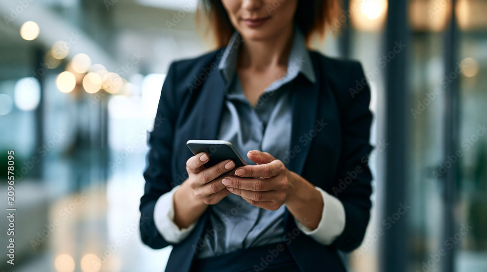 Fototapeta premium Faceless professional woman in business attire using a smartphone in a modern office corridor, quick decision-making and communication in a corporate lifestyle, defocused office