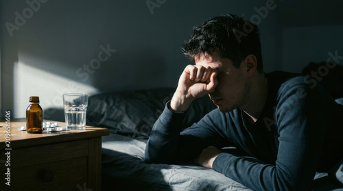 Man In Dark Room Experiencing Fatigue And Illness With Medication Nearby