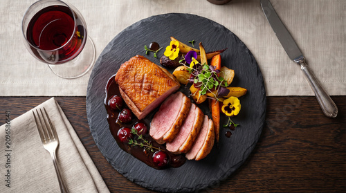 Roasted duck breast with sweet cherry reduction, beautifully paired with a crystal glass of Pinot Noir red wine on a dark slate plate in an elite fine dining restaurant.