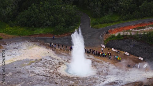 Aerial shot shows Strokkur geyser erupting in Haukadalur, Iceland. White steam rises from a turquoise pool, visitors line the vent, paths and fences encircle it.