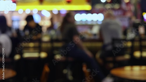 Blurred background of a busy bar or pub with customers sitting on stools at the counter. Soft bokeh lights and out of focus silhouettes of people enjoying nightlife, dining and drinking.