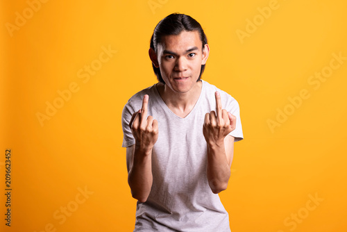Картината върху платно Irritated young man reacts with anger and defiance against yellow background, flipping both middle fingers at camera in rebellious attitude