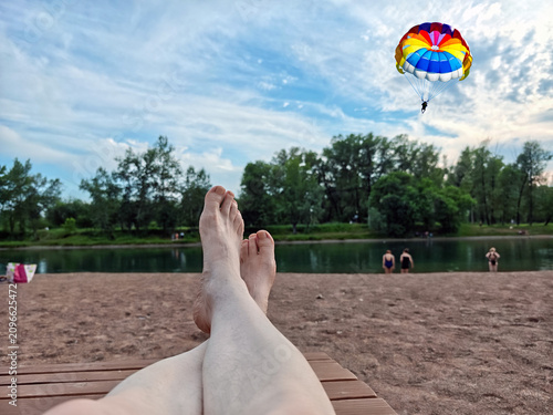 Relaxing scene of people relaxing on a sandy beach, with a view of the river and a paraglider in the sky.
