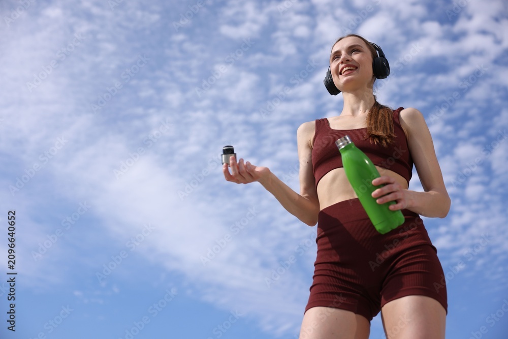 Obraz premium Woman in sportswear with headphones and water bottle outdoors under blue sky, low angle view. Space for text