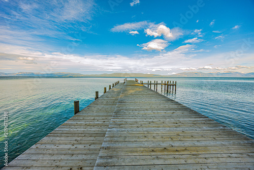Long wooden dock extending into crystal-clear turquoise waters of Lake Tahoe with distant mountains under blue sky with clouds.