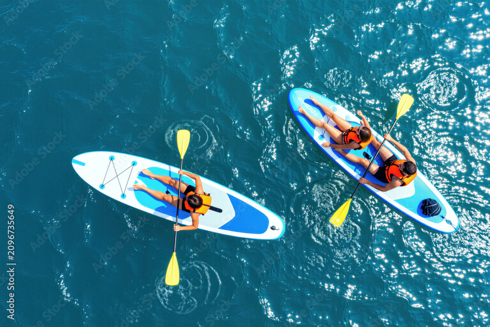 Fototapeta premium Couple paddle boards on clear blue water. People enjoy summer day on sup boards wearing life vests. They relax together during vacation on calm ocean.