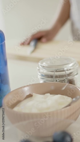 Vertical video: Framing yogurt bowl on counter, adult arm entering right cutting to add blueberries