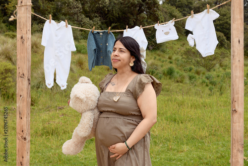 Medium Shot of Pregnant Hispanic Woman Looking Away with Teddy Bear and Baby Clothesline