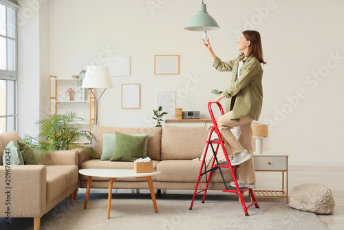 Canvastavla Beautiful young woman on stepladder changing light bulb in hanging lamp at home