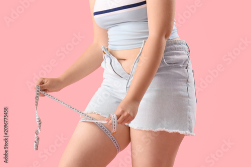 Young woman in tight shorts measuring her thigh on pink background, closeup. ...