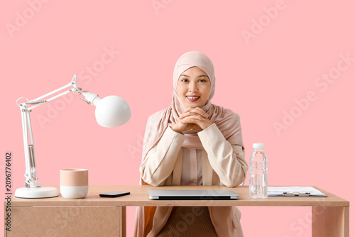 Young Muslim businesswoman at table on pink background