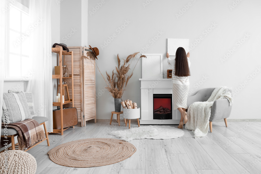 Obraz premium Young woman hanging painting on light wall above fireplace at home, back view