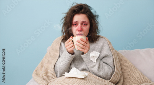 Sick young woman with red eyes and nose holding a steaming mug of hot tea wrapped in a beige blanket, feeling unwell with a cold or flu