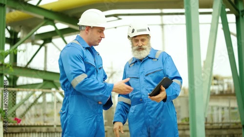 Two male industrial engineers in blue coveralls and hard hats shake hands and discuss project details on a digital tablet at an outdoor manufacturing plant, highlighting teamwork and safety.