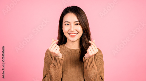 Lovely young Asian woman in a brown shirt making a finger heart gestures, isolated pink background