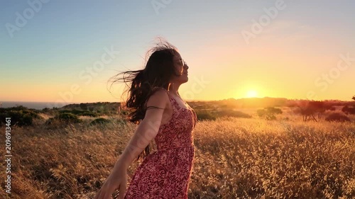 A young woman stands with arms outstretched, feeling happy and free in a beautiful golden field as the sun sets. Her long hair blows gently in the wind, embracing the peaceful nature.