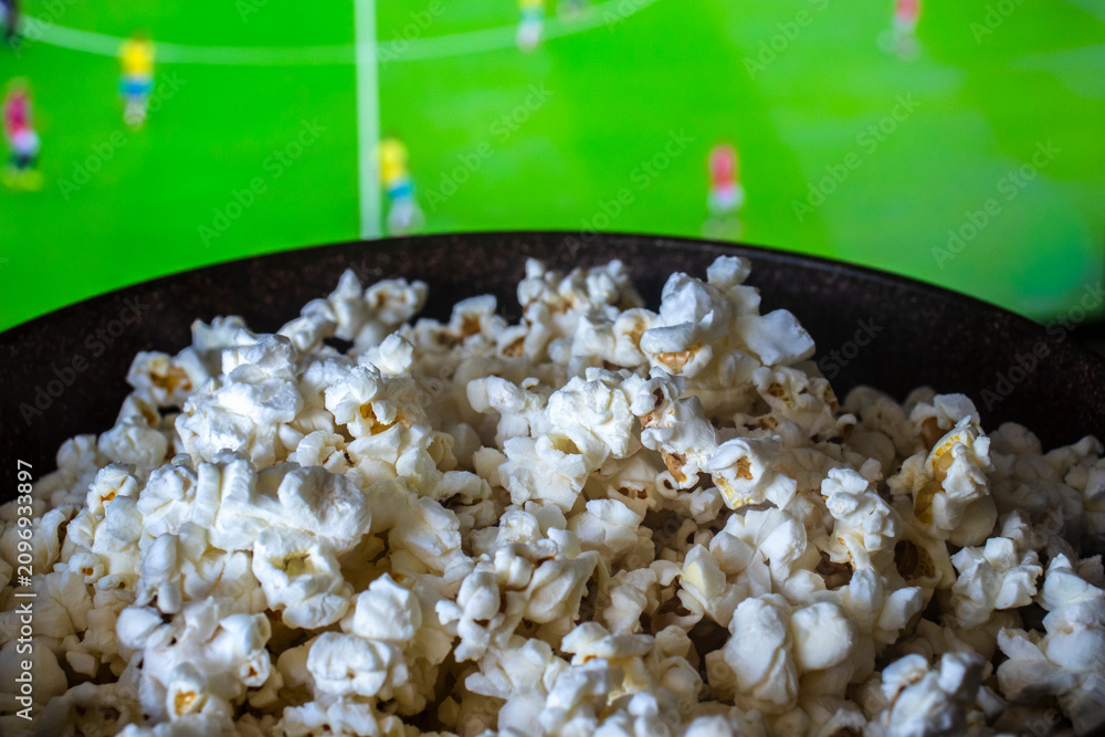 Obraz premium A delicious bowl of salty popcorn in focus with a green football field showing an active sports game blurred on a television screen at home in Brazil.