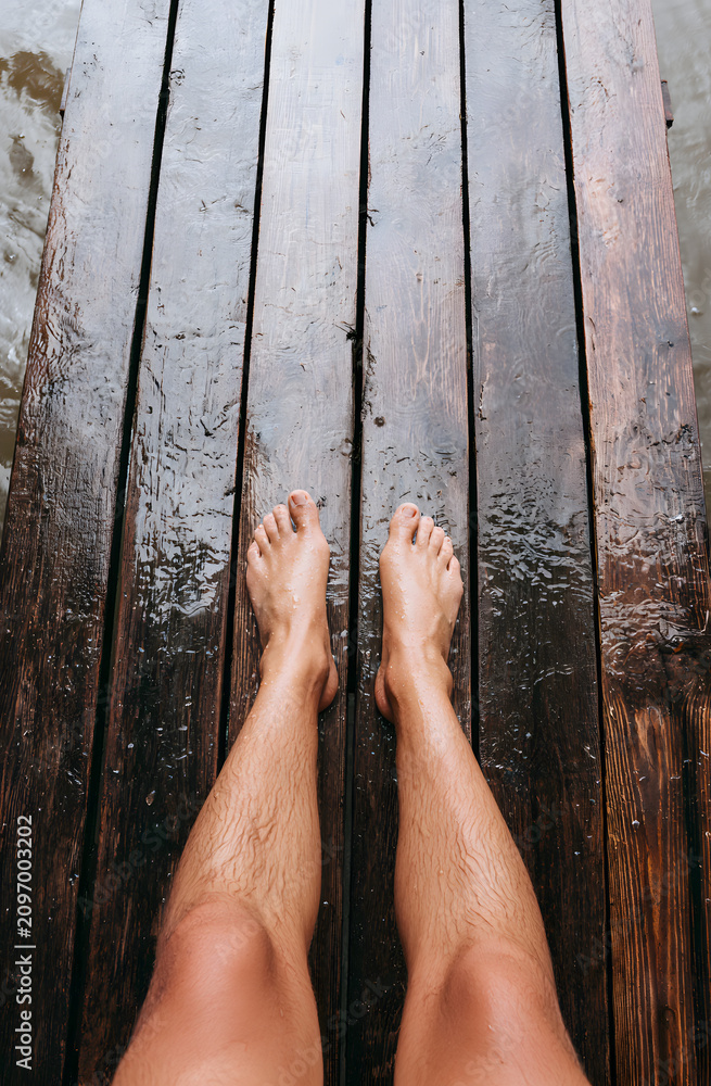 Fototapeta premium Unshod feet rest on a soggy wooden boardwalk. Two individuals' legs, with one knee lower than the other, skim wet planks near the water. Summertime leisure by the pier.