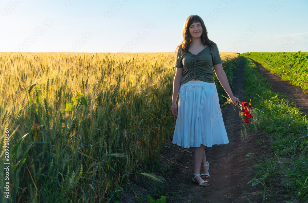 Obraz premium Young woman walking in wheat field holding red flowers at sunset