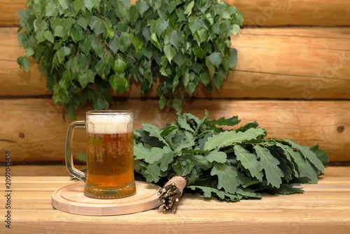 Foto A mug of light beer on a round wooden tray next to a fresh oak broom stands on a plank bench against the background of a log wall with a couple of fresh birch brooms hang on it
