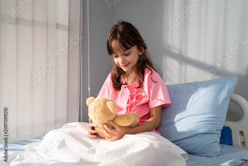Caucasian young girl sitting on a hospital bed, holding her teddy bear. 