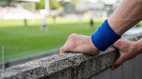 Close-up of a person's hands resting on a textured concrete wall with a blue sweatband on the wrist, overlooking a blurred professional soccer stadium with players on a green pitch.