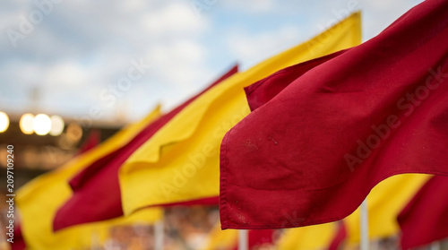 Close-up view of red and yellow fabric flags arranged in a row at a sports stadium, waving against a blue sky background with stadium lights and shallow depth of field focus.