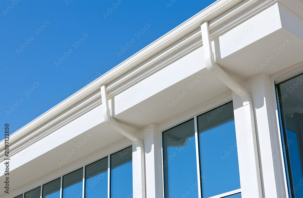 Obraz premium White rain gutters on contemporary commercial edifice. Sleek architectural feature contrasting with azure sky. Glazed surface with expansive glass panes mirrors the heavens.
