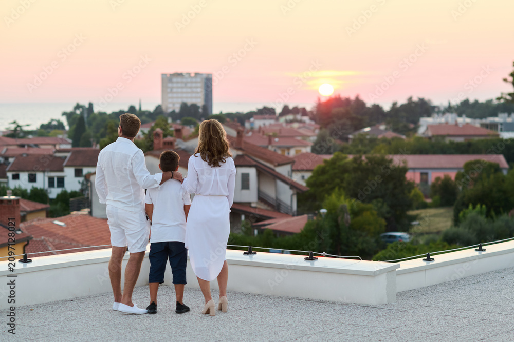 Obraz premium Family On Rooftop Terrace Watching Sunset Over Coastal Town And Sea View
