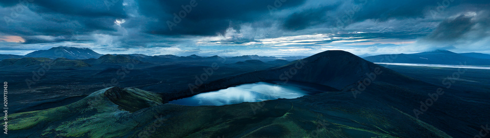 Fototapeta premium Panoramic view of Ljótipollur crater lake in Iceland's Highlands, surrounded by dark volcanic slopes beneath dramatic storm clouds. A remote and breathtaking volcanic landscape.