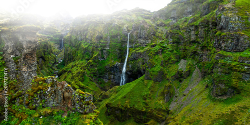 Panoramic view of the breathtaking Múlagljúfur Canyon in southeast Iceland, featuring dramatic moss-covered cliffs, a cascading waterfall, and a winding river beneath soft mountain light.
