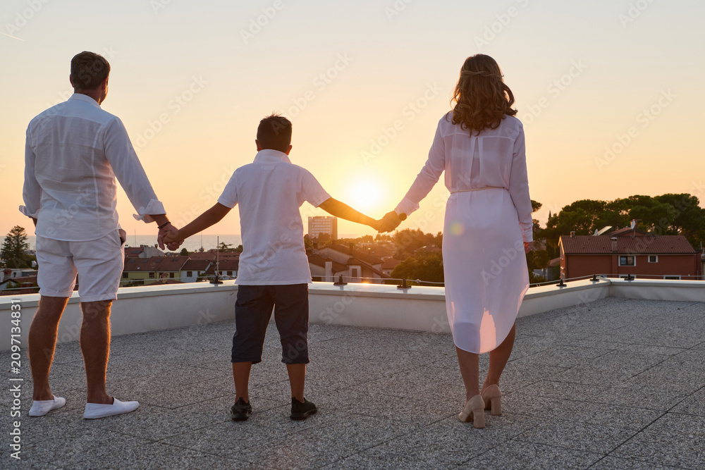 Obraz premium Family Holding Hands on Rooftop Terrace at Sunset With Sea View and Coastal Town Background