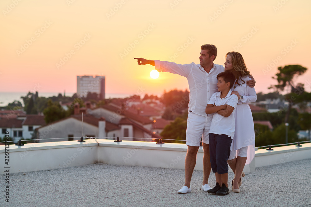 Obraz premium Family on Rooftop Balcony at Sunset With Sea View, Parents Pointing Toward Horizon