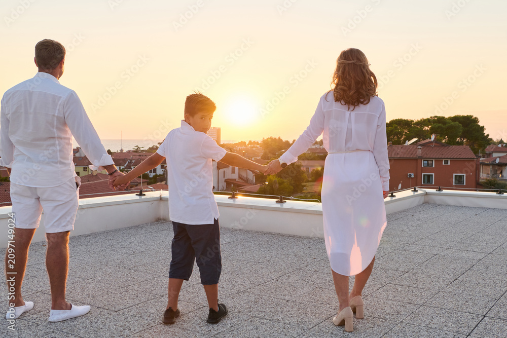 Obraz premium Family Holding Hands on Rooftop Terrace at Sunset With Sea View and Summer Vacation Moment