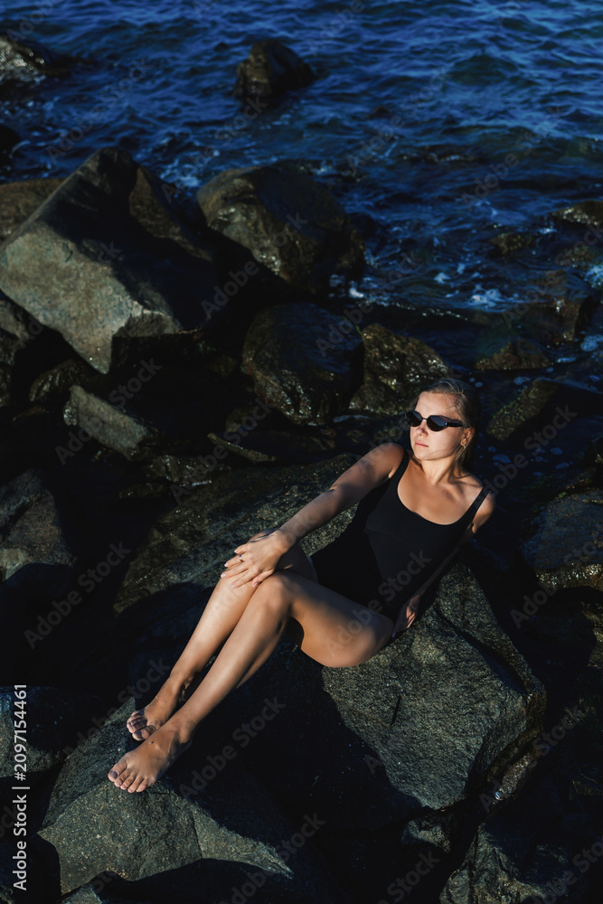 Fototapeta premium Woman relaxing on rocks by the water during daylight, wearing black swimwear and sunglasses while enjoying the outdoor setting