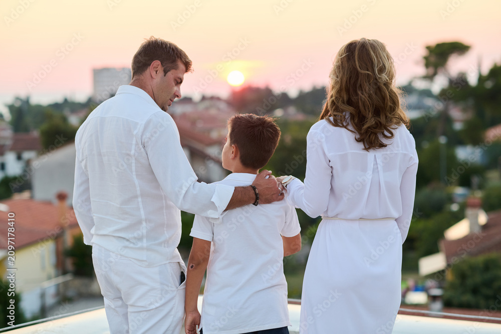 Obraz premium Family on Rooftop Balcony Watching Sunset Over Coastal Town Parents and Son Embracing Togetherness
