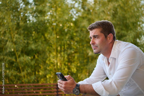 Fotografie Thoughtful Man on Balcony Holding Phone Relaxing at Home Terrace Overlooking Gre
