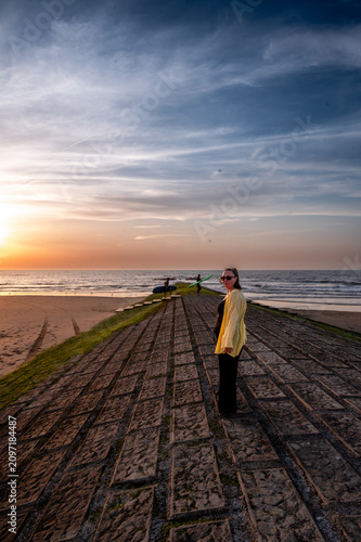 Cuadro en lienzo Woman in a yellow cardigan glancing back over her shoulder while standing on a stone breakwater at sunset a calm and reflective coastal moment in warm summer evening light